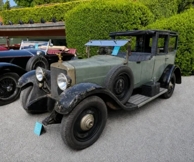 Ein historischer, grau-schwarzer Vorkriegs-Oldtimer mit großen verchromten Rundscheinwerfern und einem seitlich montierten Ersatzrad steht ausgestellt neben weiteren klassischen Fahrzeugen auf einem asphaltierten Platz vor einer grünen Heckenkulisse.