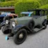 Ein historischer, grau-schwarzer Vorkriegs-Oldtimer mit großen verchromten Rundscheinwerfern und einem seitlich montierten Ersatzrad steht ausgestellt neben weiteren klassischen Fahrzeugen auf einem asphaltierten Platz vor einer grünen Heckenkulisse.