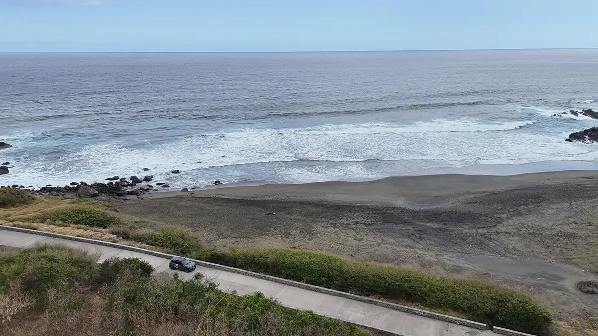 Luftaufnahme einer zweispurigen Küstenstrasse auf La Réunion, auf der ein dunkles Fahrzeug direkt am wilden, blau schäumenden Indischen Ozean entlangfährt.