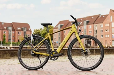 Ein schlankes, gold-olivgrünes Hercules Urbanico E-Bike mit Diamantrahmen, Vollausstattung und Gepäckträger vor einer städtischen Kulisse mit Backsteingebäuden.