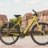 Ein schlankes, gold-olivgrünes Hercules Urbanico E-Bike mit Diamantrahmen, Vollausstattung und Gepäckträger vor einer städtischen Kulisse mit Backsteingebäuden.