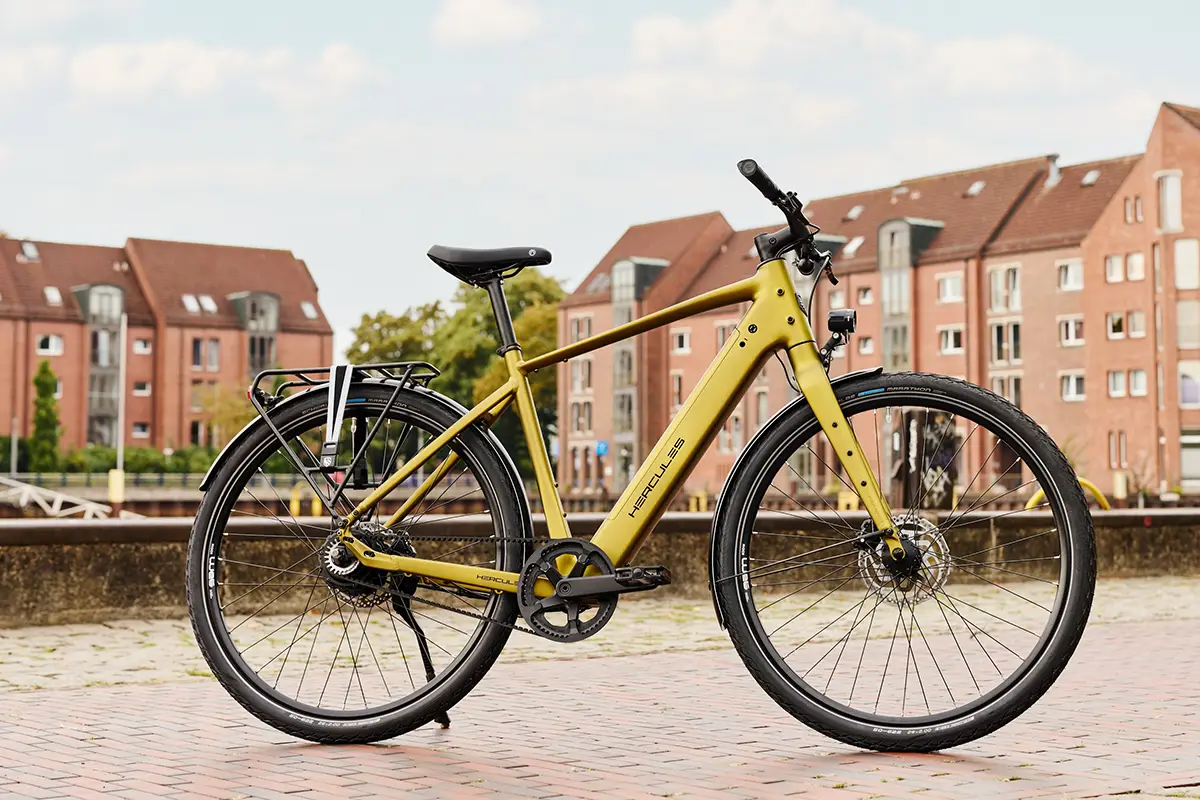 Ein schlankes, gold-olivgrünes Hercules Urbanico E-Bike mit Diamantrahmen, Vollausstattung und Gepäckträger vor einer städtischen Kulisse mit Backsteingebäuden.