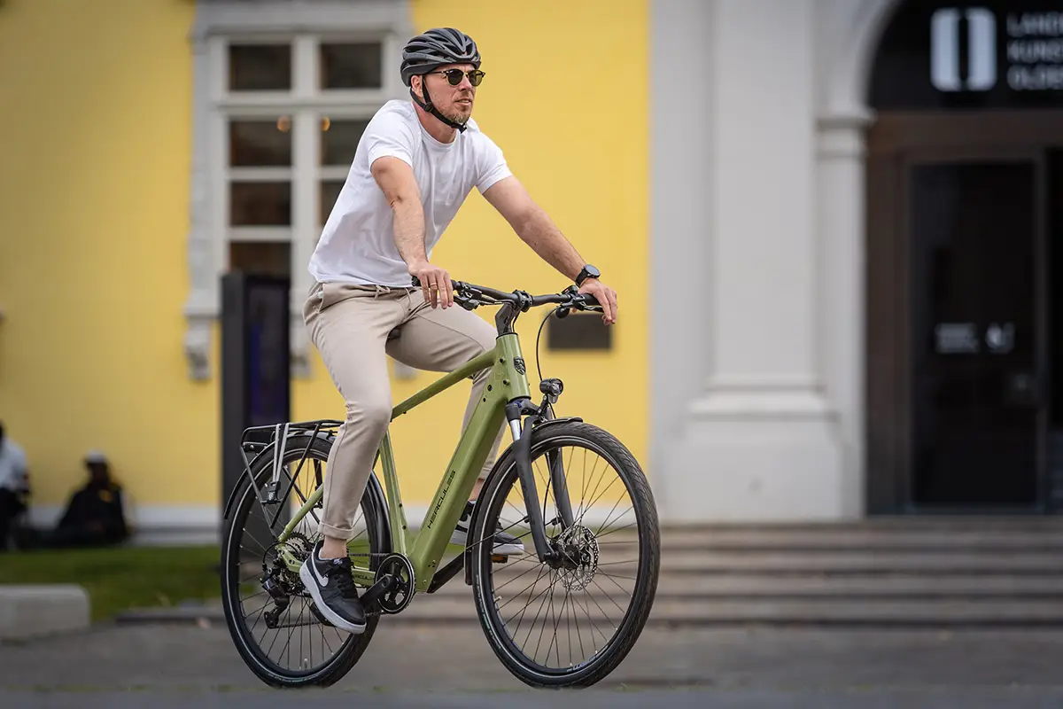 Eine Person mit Fahrradhelm und Sonnenbrille fährt auf dem olivgrünen Hercules Urbanico E-Bike mit Kettenschaltung auf einer asphaltierten Straße vor einem gelben Gebäude.