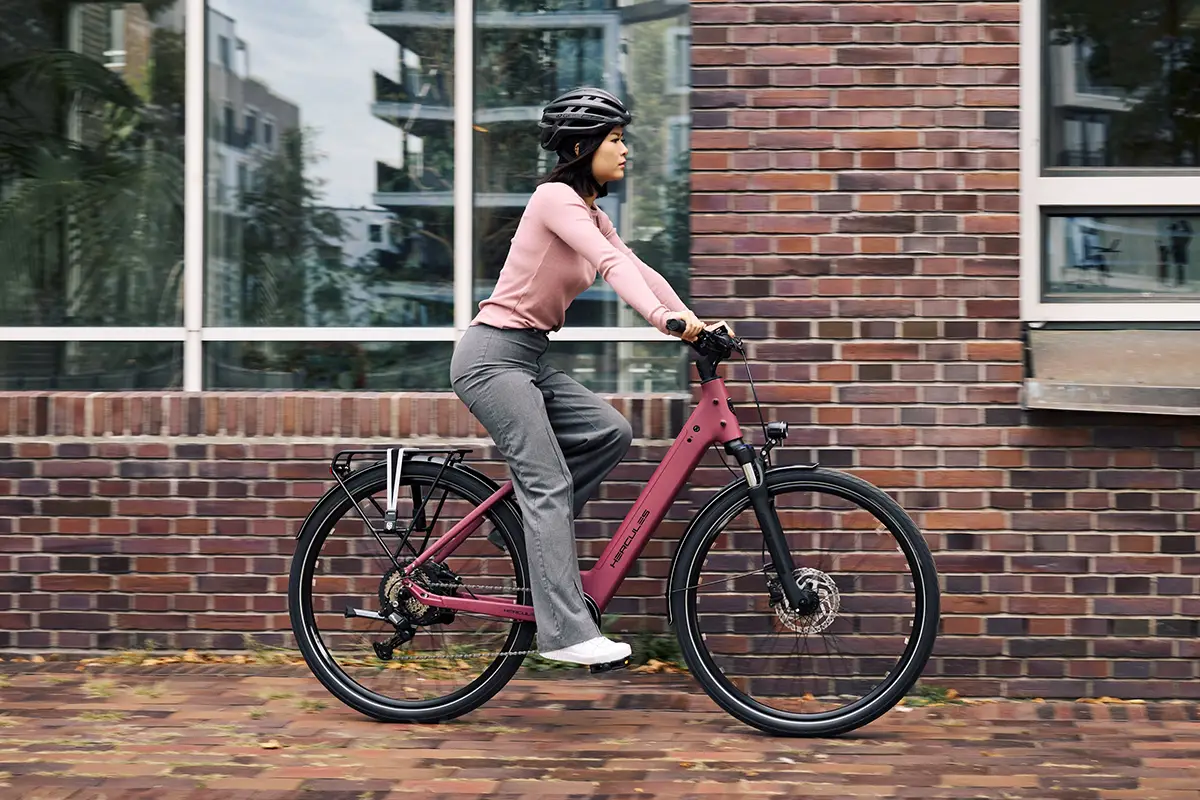 Eine Person mit Fahrradhelm fährt auf einem beerenroten Hercules Urbanico E-Bike mit Trapezrahmen entlang einer modernen Backstein-Häuserfassade.