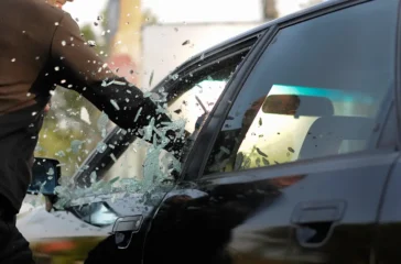 Nahaufnahme einer geschützten Hand, die im Notfall mit einem spitzen Gegenstand nahe am Rand ansetzt und die Seitenscheibe eines dunklen Fahrzeugs einschlägt, während das Autoglas splittert.