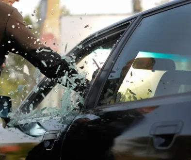 Nahaufnahme einer geschützten Hand, die im Notfall mit einem spitzen Gegenstand nahe am Rand ansetzt und die Seitenscheibe eines dunklen Fahrzeugs einschlägt, während das Autoglas splittert.