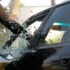 Nahaufnahme einer geschützten Hand, die im Notfall mit einem spitzen Gegenstand nahe am Rand ansetzt und die Seitenscheibe eines dunklen Fahrzeugs einschlägt, während das Autoglas splittert.