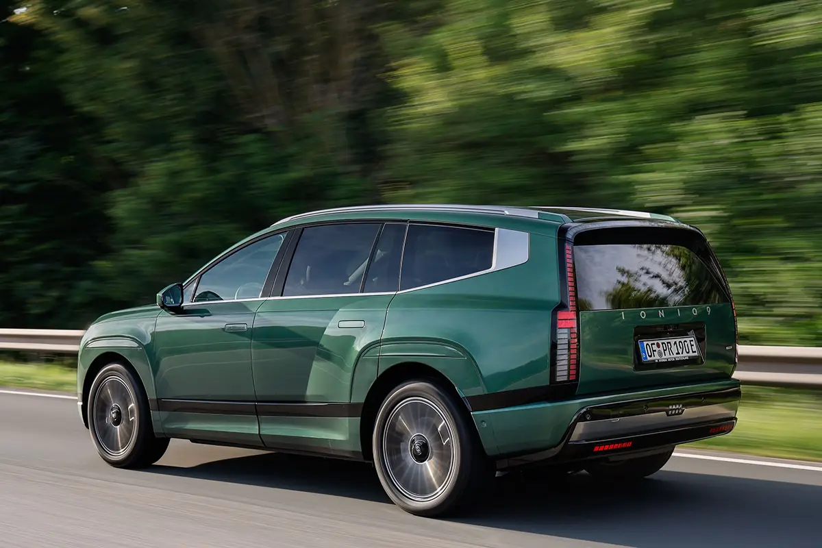 Heckperspektive des grossen, dunkelgrünen Elektro-SUVs Hyundai Ioniq 9 während der Fahrt auf einer asphaltierten Autobahn vor einer Kulisse aus grünen Bäumen.