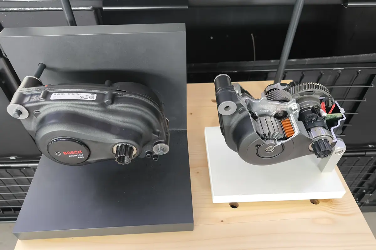 Produkt-Präsentation des neuen Bosch Active Line Plus E-Bike-Mittelmotors; links das geschlossene, kompakte Serienaggregat und rechts ein aufgeschnittenes Gehäusemodell, das die interne Getriebestruktur und den Elektromotor zeigt.