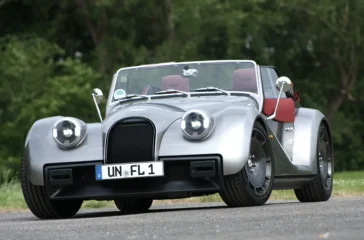 Frontansicht des silbernen Morgan Supersport Roadsters mit geöffnetem Verdeck und rotem Interieur auf einer Landstraße.