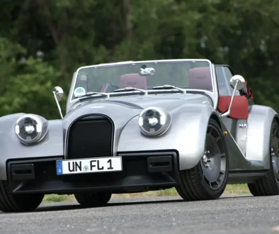 Frontansicht des silbernen Morgan Supersport Roadsters mit geöffnetem Verdeck und rotem Interieur auf einer Landstraße.