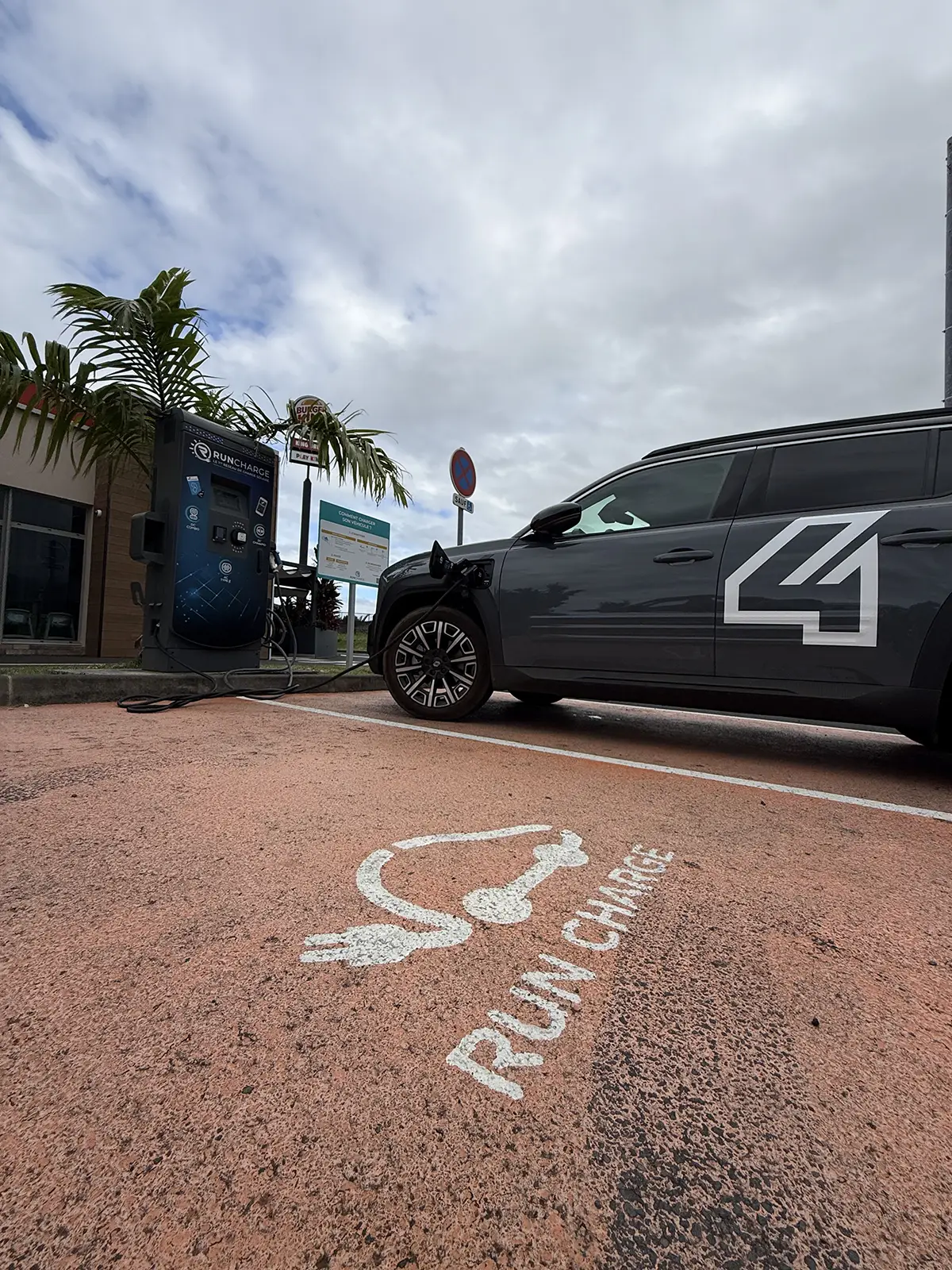 Nahaufnahme der rechten Fahrzeugseite des grauen Renault 4 Elektro an einer Ladesäule auf einem Parkplatz mit der Bodenmarkierung RUN CHARGE unter bewölktem Himmel