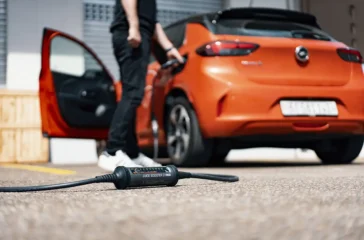 Heckseitenansicht eines orangefarbenen Elektroautos mit geöffneter Fahrertür, bei dem ein mobiles Ladegerät (Juice Booster) am Ladeanschluss angeschlossen ist, während das Steuergerät flach auf dem Asphalt liegt.