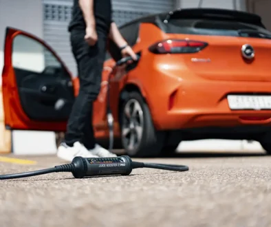 Heckseitenansicht eines orangefarbenen Elektroautos mit geöffneter Fahrertür, bei dem ein mobiles Ladegerät (Juice Booster) am Ladeanschluss angeschlossen ist, während das Steuergerät flach auf dem Asphalt liegt.