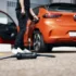 Heckseitenansicht eines orangefarbenen Elektroautos mit geöffneter Fahrertür, bei dem ein mobiles Ladegerät (Juice Booster) am Ladeanschluss angeschlossen ist, während das Steuergerät flach auf dem Asphalt liegt.