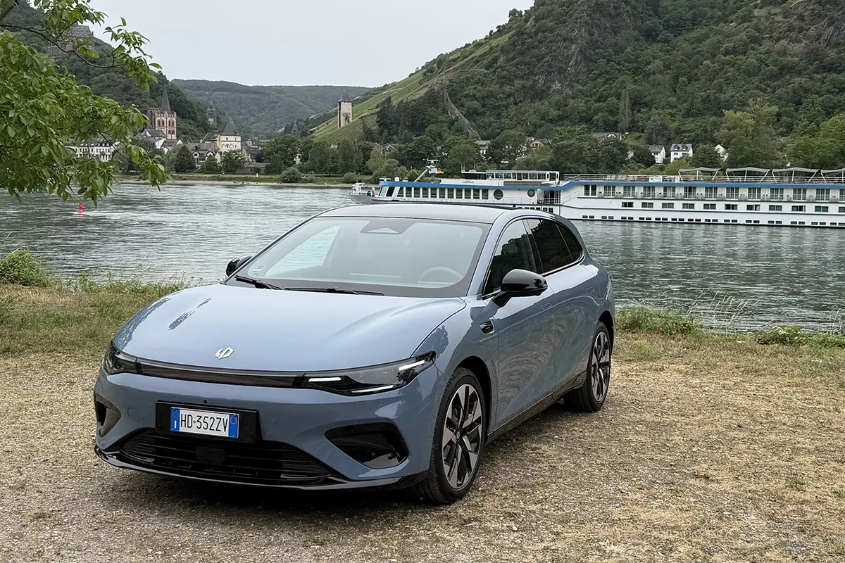 Schrägansicht von vorne auf das blaugraue Elektro-Kompakt-SUV Leapmotor B05, das auf kieseligem Untergrund am Ufer einer Flusslandschaft vor bewaldeten Hügeln steht.