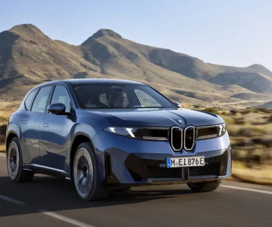 Ein moderner, tiefblauer BMW iX3 Elektro-Crossover aus der zukunftsweisenden „Neuen Klasse“ in dynamischer Fahrt auf einer asphaltierten Landstraße vor einer weitläufigen, trockenen Wüsten- und Bergkulisse.