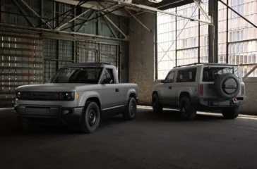 Der graue, unlackierte Slate Elektro-Pick-up als kompakter Zweitürer (links) und die längere, fünfsitzige Slate SUV-Variante mit aussen am Heck montiertem Ersatzrad (rechts) parken nebeneinander in einer historischen, hellen Industriehalle.