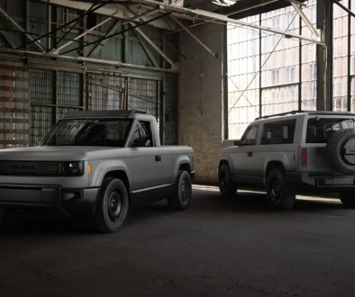 Der graue, unlackierte Slate Elektro-Pick-up als kompakter Zweitürer (links) und die längere, fünfsitzige Slate SUV-Variante mit aussen am Heck montiertem Ersatzrad (rechts) parken nebeneinander in einer historischen, hellen Industriehalle.