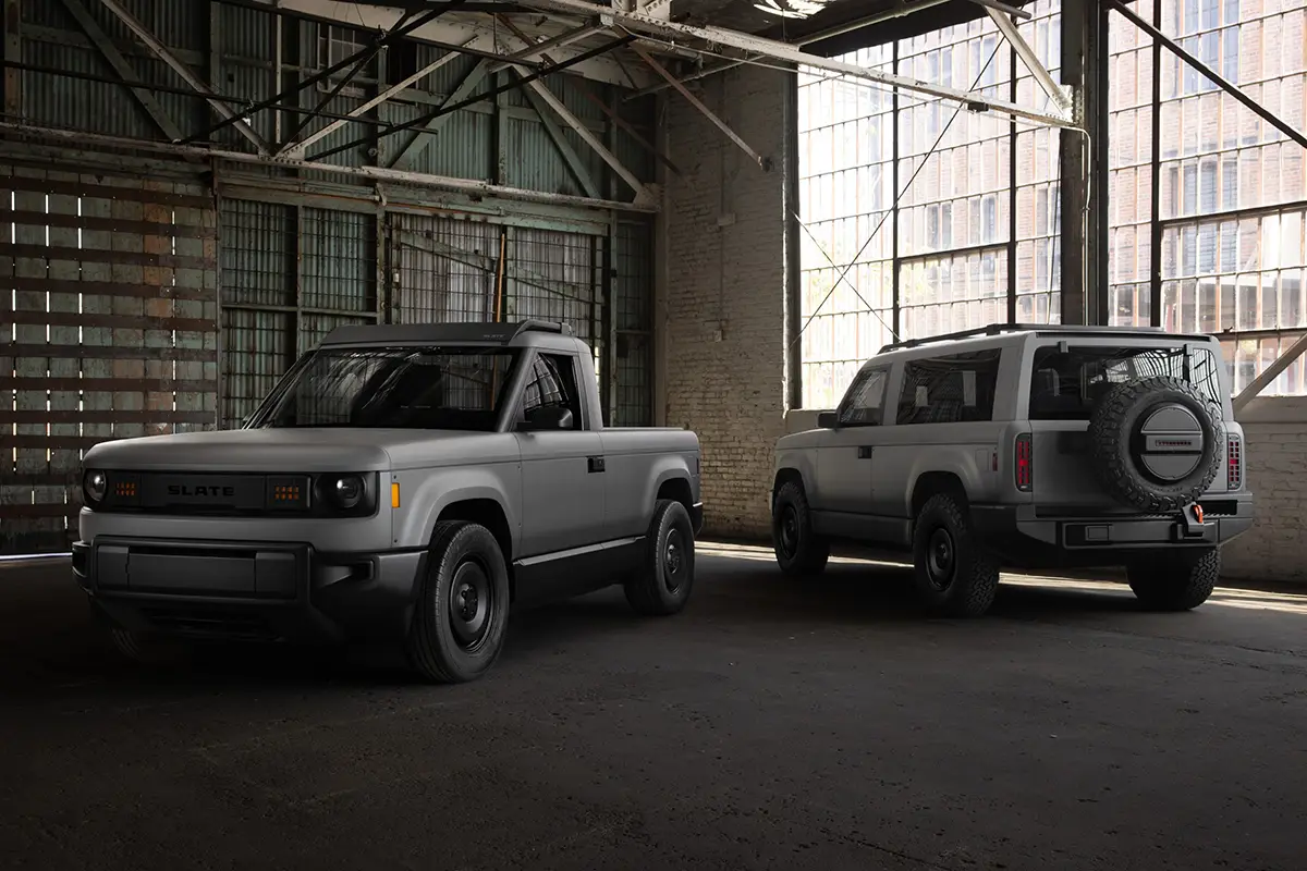 Der graue, unlackierte Slate Elektro-Pick-up als kompakter Zweitürer (links) und die längere, fünfsitzige Slate SUV-Variante mit aussen am Heck montiertem Ersatzrad (rechts) parken nebeneinander in einer historischen, hellen Industriehalle.