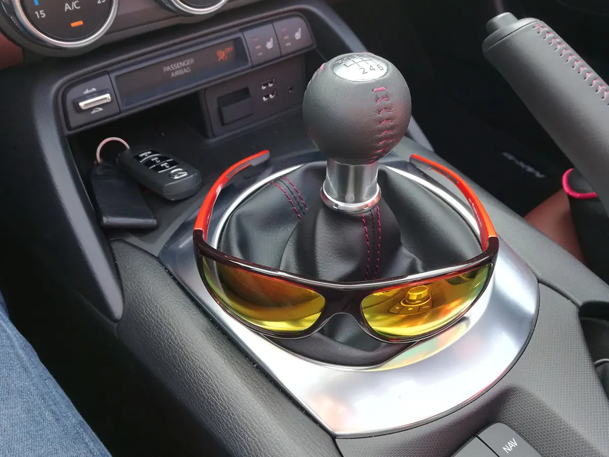 Nahaufnahme des fahrzeuginternen Cockpits im Sommer mit einer sportlichen, orange-gelb verspiegelten Sonnenbrille, die über der Leder-Manschette des manuellen Schalthebels abgelegt wurde.