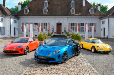 Drei Generationen von Alpine-Sportwagen – ein moderner orangefarbener A110, eine blau-schwarz lackierte A110 R Ultime und ein klassischer gelber Oldtimer – stehen nebeneinander auf dem gepflasterten Vorplatz des barocken Schlosses Thunstetten.