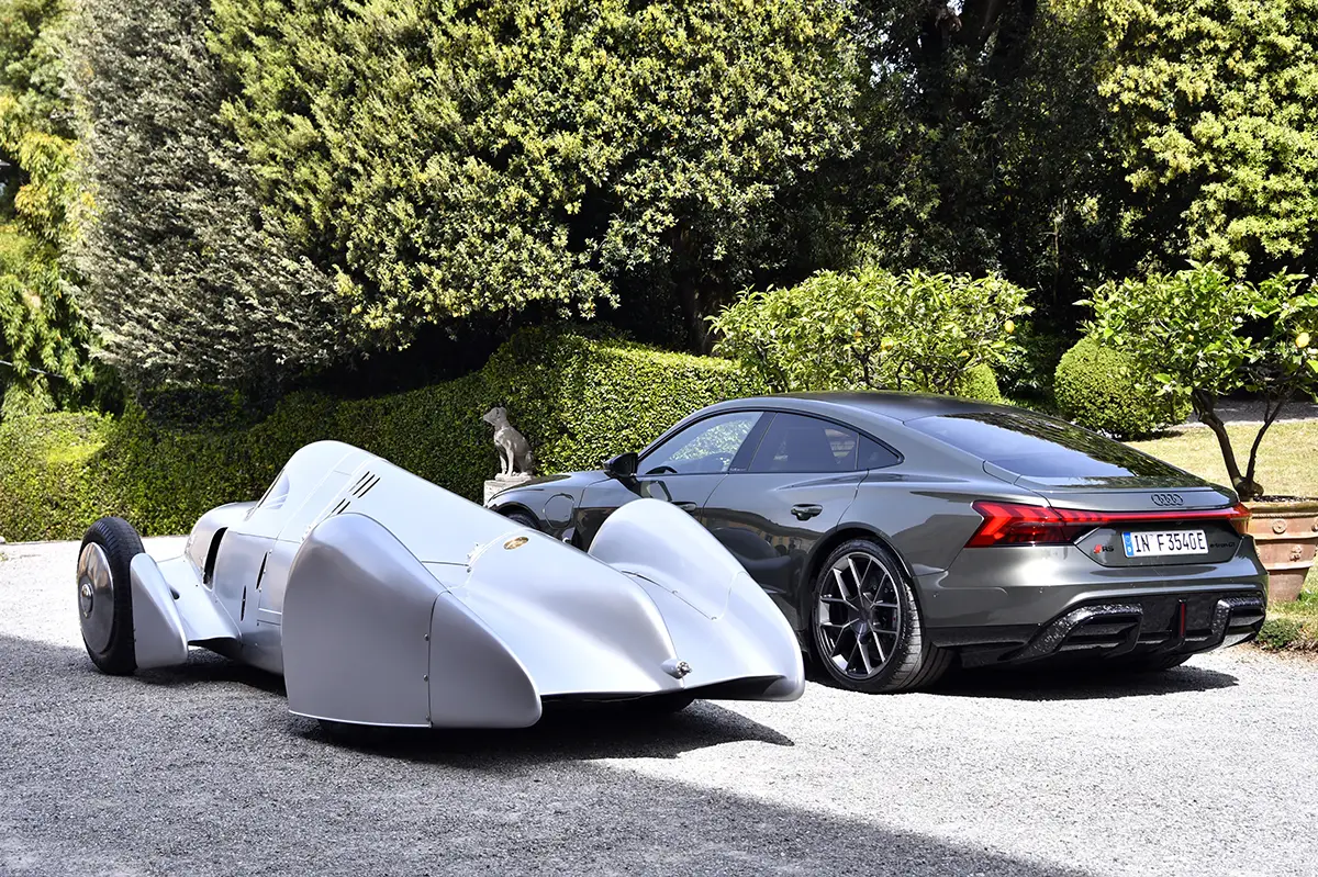 Der extrem flache und aerodynamische Auto Union Typ A Silberpfeil parkt im Schatten grosser Bäume neben dem kantigen Heck eines anthrazitfarbenen Audi RS e-tron GT performance.