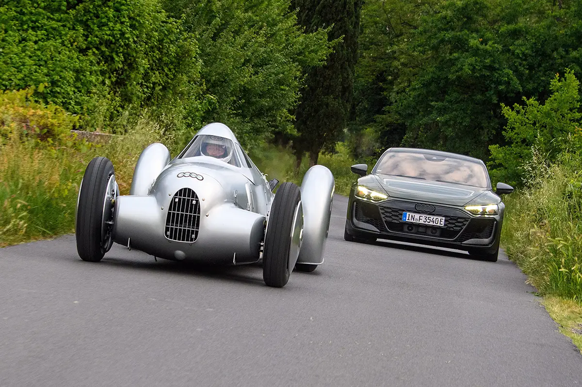 Der historische, silberne Auto Union Typ A Stromlinienwagen „Lucca“ fährt auf einer schmalen Asphaltstrasse in der Toskana, dicht gefolgt von einem modernen, dunkelgrauen Audi RS e-tron GT.