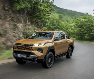 Ein gold-bronze-farbener Toyota Hilux der neunten Generation fährt dynamisch auf einer asphaltierten Straße entlang einer bewaldeten Hügellandschaft.