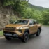Ein gold-bronze-farbener Toyota Hilux der neunten Generation fährt dynamisch auf einer asphaltierten Straße entlang einer bewaldeten Hügellandschaft.