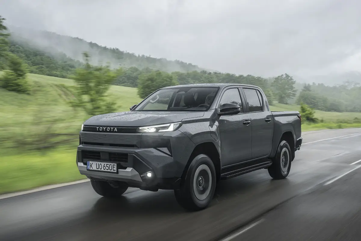 Seitenprofil des vollelektrischen Toyota Hilux Elektro-Pickups in Dunkelgrau während der Fahrt vor grünem Hintergrund.