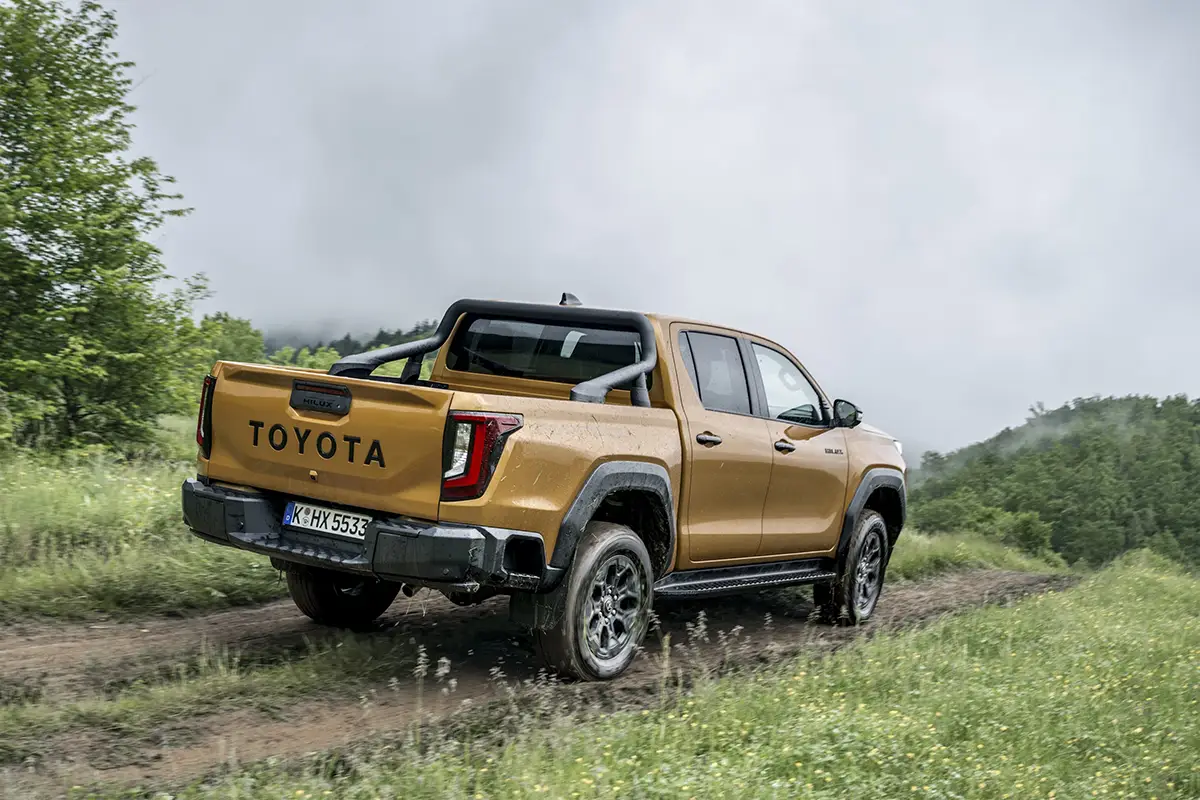 Die Heckansicht des gold-bronze-farbenen Toyota Hilux Doppelkabine mit dem großen, eingeprägten TOYOTA-Schriftzug auf der Ladeklappe im Gelände.