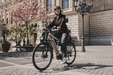 Eine E-Bikerin fährt im urbanen Raum auf einem Pflastersteinweg vor einem historischen Steingebäude in Hamburg mit einem City-E-Bike, das mit dem neuen Bosch Active Line Plus Smart System ausgestattet ist.