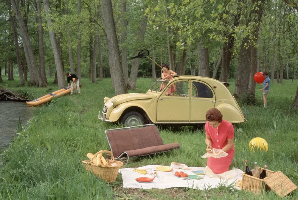 Forest picnic near a yellow vintage car; a woman lays out food on a blanket with baskets nearby.