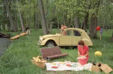 Forest picnic near a yellow vintage car; a woman lays out food on a blanket with baskets nearby.