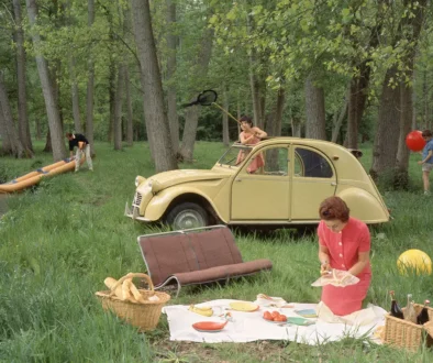 Forest picnic near a yellow vintage car; a woman lays out food on a blanket with baskets nearby.