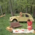 Forest picnic near a yellow vintage car; a woman lays out food on a blanket with baskets nearby.