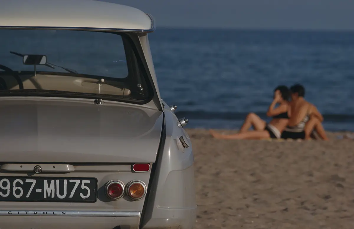 Rear view of a vintage Citroën on a sandy beach, with a couple sunbathing near the calm sea in the background.