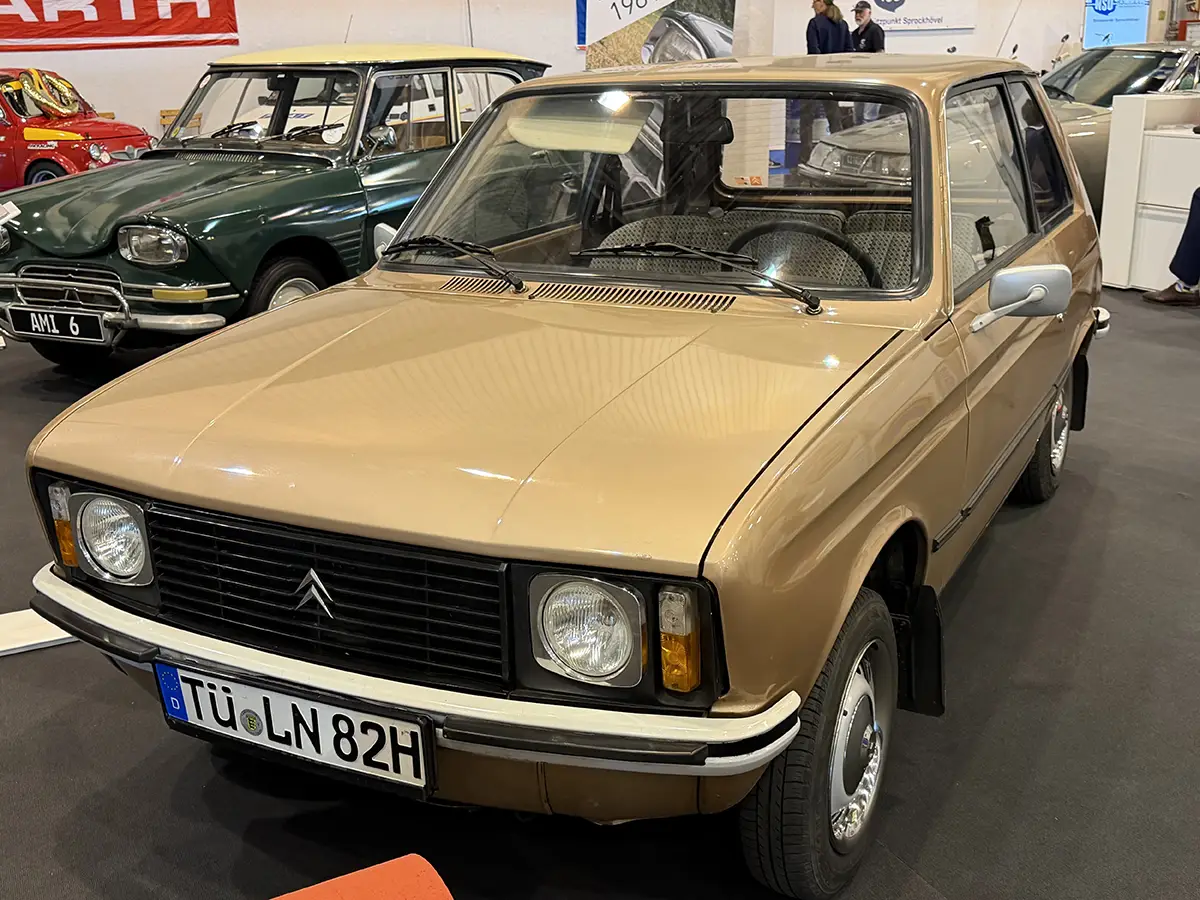 Beige vintage hatchback car on a display floor at a classic car show, front view with black grille and round headlights (license plate visible).