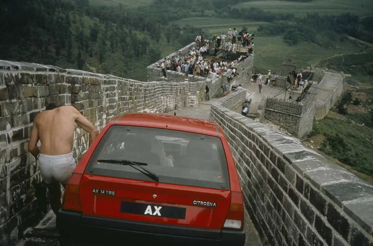 Shirtless man beside a red hatchback parked on a narrow Great Wall segment with crowds walking along the stone path nearby.