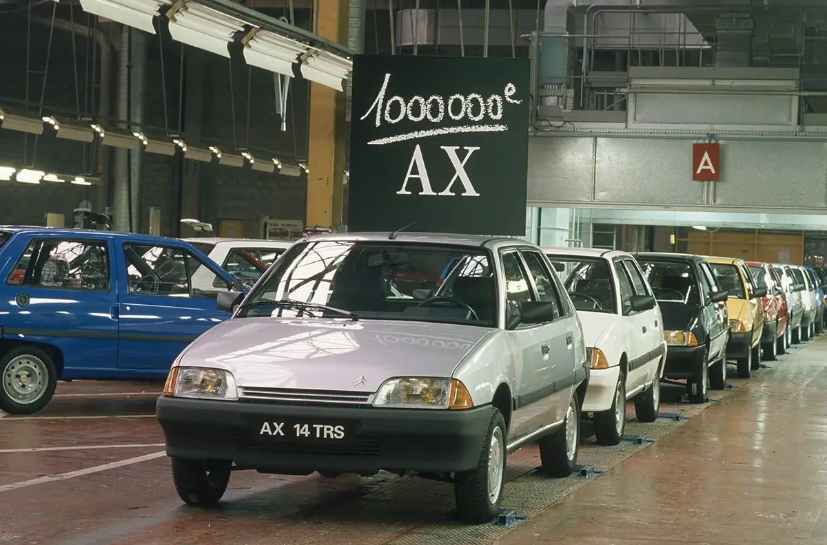 Line of compact cars on an automotive assembly floor; silver car in front shows AX 14 TRS license plate