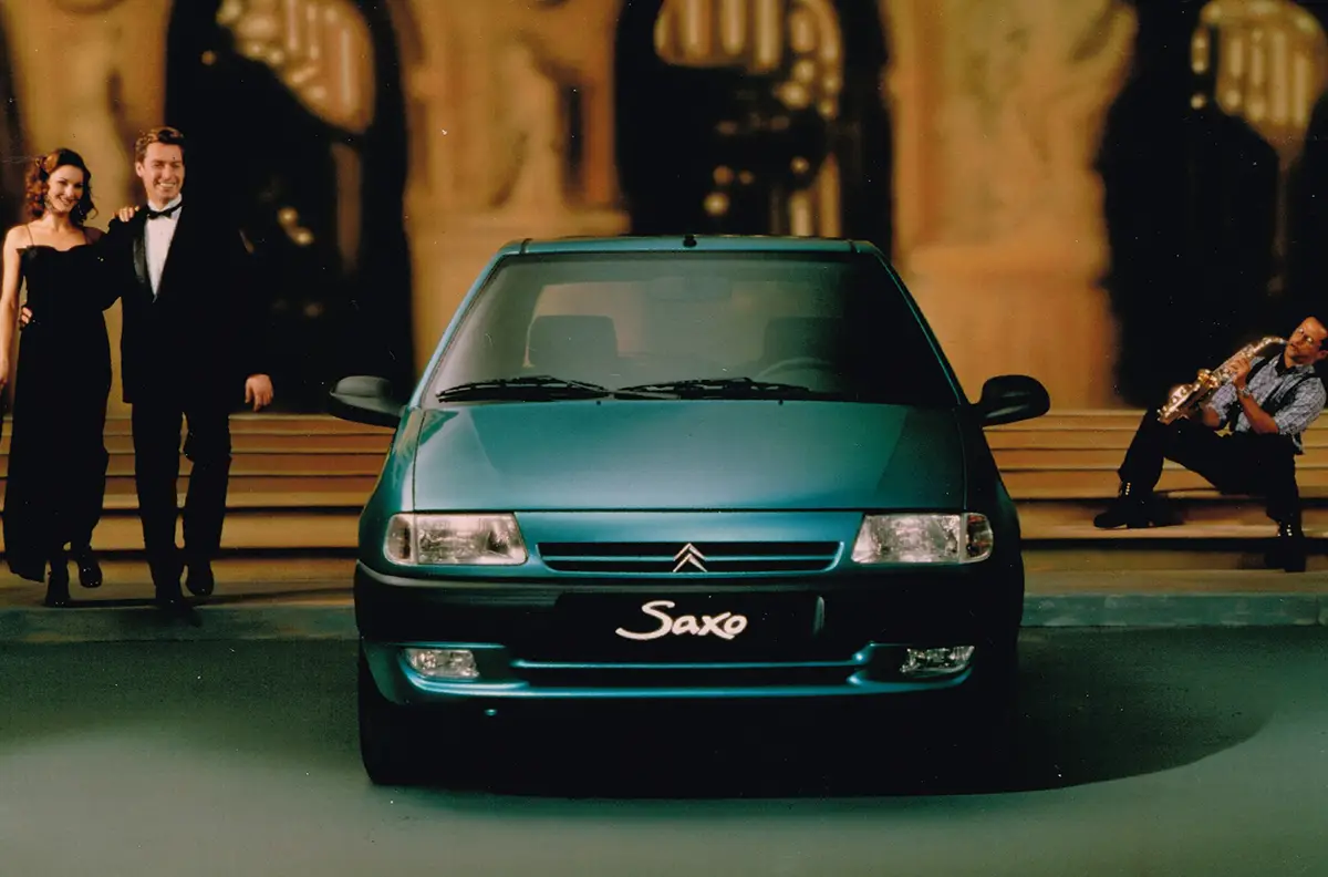 Teal Citroën Saxo parked center with a formally dressed couple on the left and a saxophone player on the right. The scene resembles a night event on steps behind the car, with 'Saxo' visible on the grille.