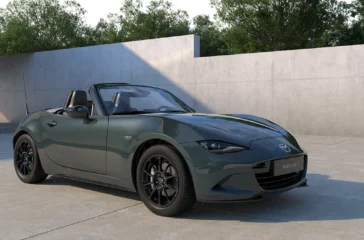 Ein offener Mazda MX-5 Roadster des Modelljahrs 2027 mit der neuen, dunkelgrünen Metallic-Lackierung „Zinc Green“ und schwarzen Leichtmetallfelgen parkiert in einem minimalistischen Studio vor einer grauen Betonwand.