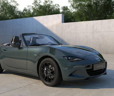Ein offener Mazda MX-5 Roadster des Modelljahrs 2027 mit der neuen, dunkelgrünen Metallic-Lackierung „Zinc Green“ und schwarzen Leichtmetallfelgen parkiert in einem minimalistischen Studio vor einer grauen Betonwand.