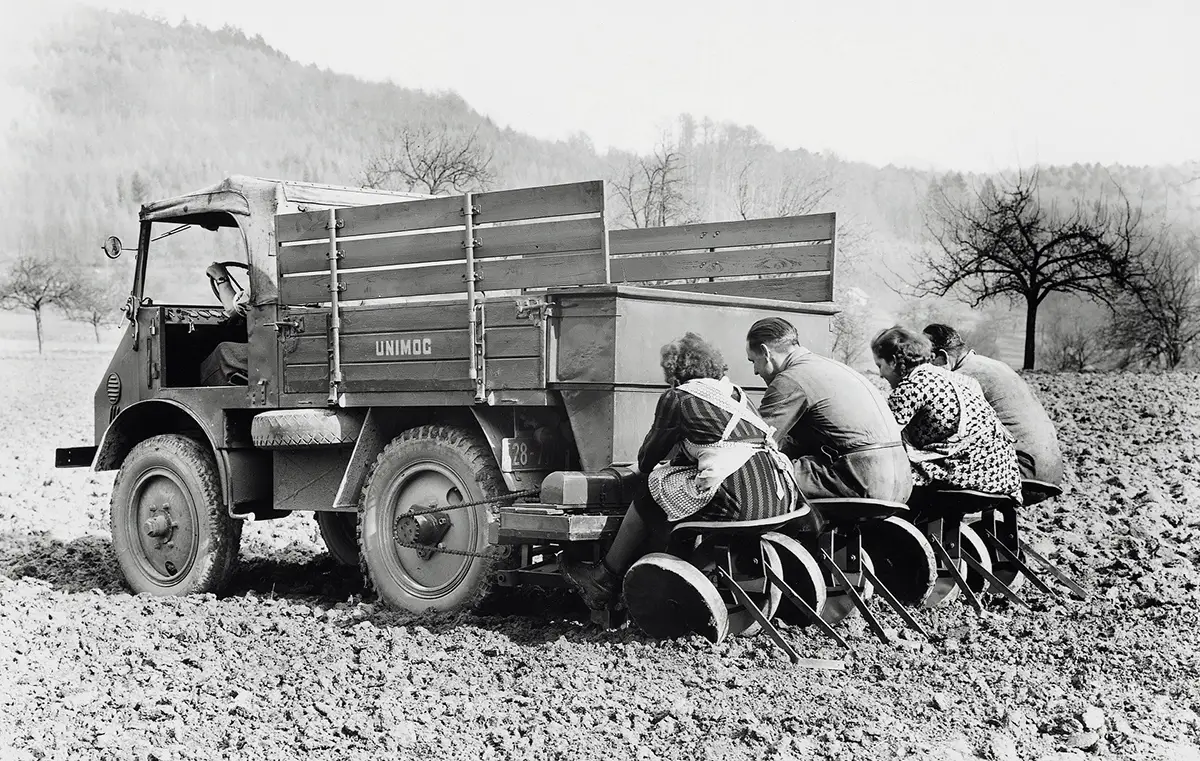 Historische Schwarz-Weiss-Aufnahme eines frühen Unimog mit Landarbeitern auf einer Kartoffelpflanzmaschine im Feld.