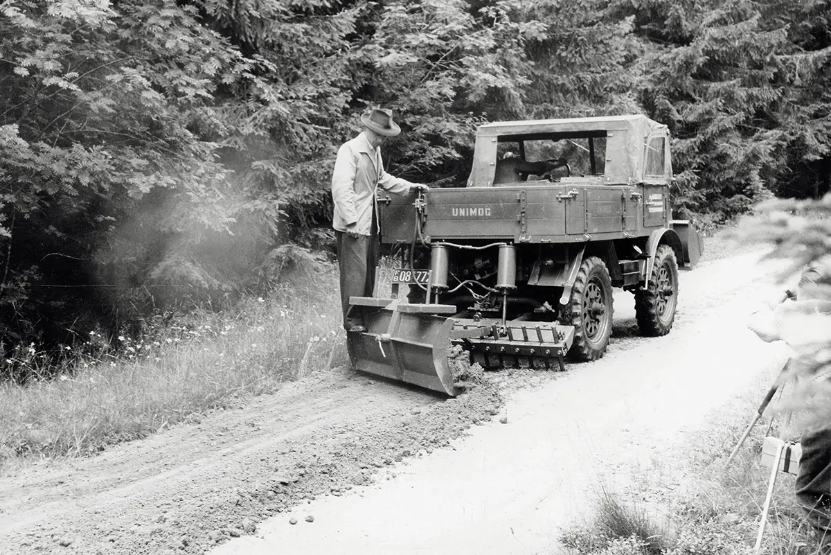 Historisches Foto eines Unimog U 25 mit Planierschild bei Wegearbeiten im Wald in den 1950er Jahren.