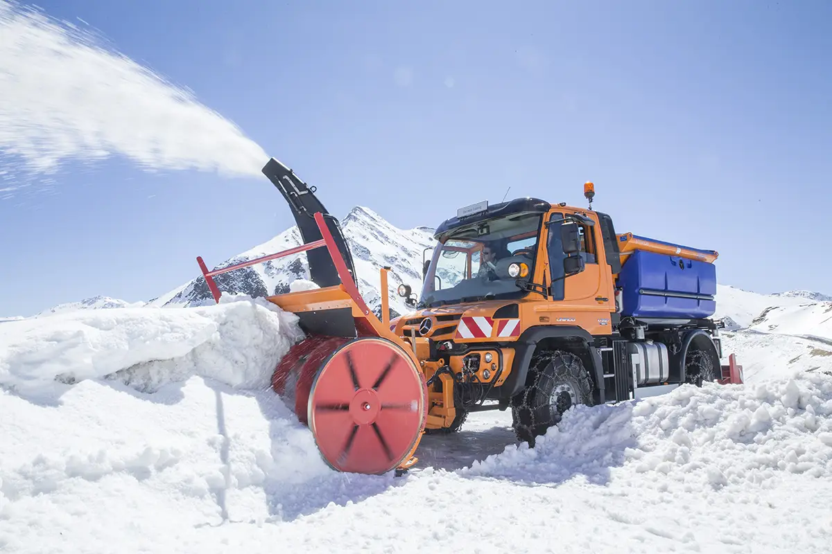 Orangefarbener Unimog mit leistungsstarker Schneefräse räumt gewaltige Schneemassen auf einer hochalpinen Gebirgsstrasse.