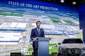 Mercedes-Benz-Produktionsvorstand Michael Schiebe [cite: 543] spricht an einem Rednerpult vor einer riesigen Leinwand mit der Aufschrift „STATE-OF-THE-ART PRODUCTION“[cite: 542], die eine Luftaufnahme des ungarischen Mercedes-Werks in Kecskemét zeigt.