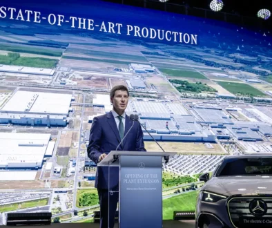Mercedes-Benz-Produktionsvorstand Michael Schiebe [cite: 543] spricht an einem Rednerpult vor einer riesigen Leinwand mit der Aufschrift „STATE-OF-THE-ART PRODUCTION“[cite: 542], die eine Luftaufnahme des ungarischen Mercedes-Werks in Kecskemét zeigt.
