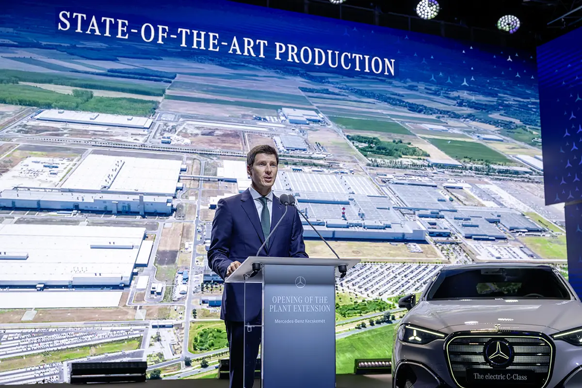 Mercedes-Benz-Produktionsvorstand Michael Schiebe [cite: 543] spricht an einem Rednerpult vor einer riesigen Leinwand mit der Aufschrift „STATE-OF-THE-ART PRODUCTION“[cite: 542], die eine Luftaufnahme des ungarischen Mercedes-Werks in Kecskemét zeigt.